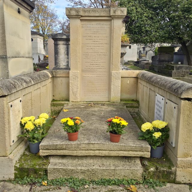 Grave of the Baugnies family with a slanted double-pitched stone slab on a base, surrounded by stone screens with inscriptions. A tall aedicula with fluted pilasters and palm leaf reliefs stands at the head. Potted flowers are placed in front.