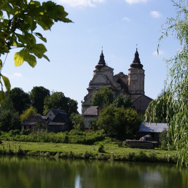 The ruins of the Church of St. Mark in Waręż, surrounded by trees and located near a pond, with a clear blue sky in the background.