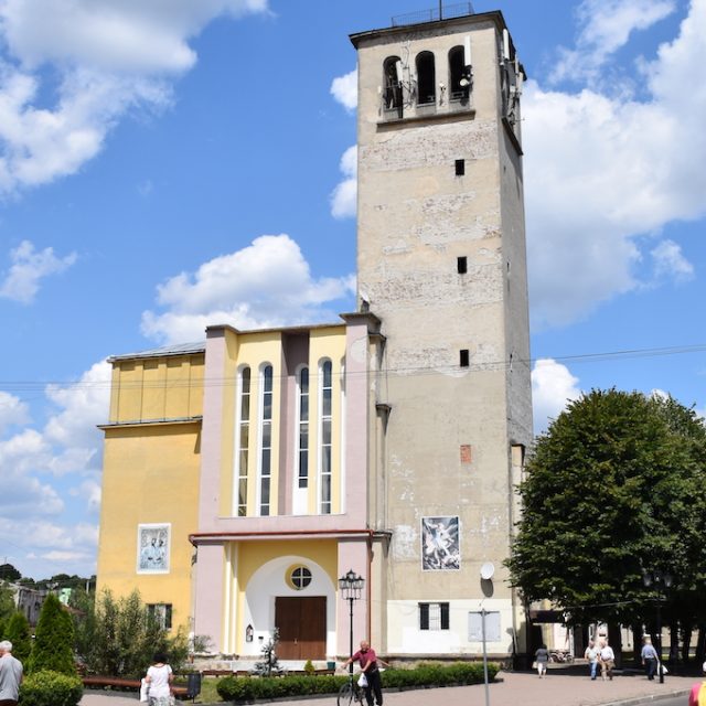 L'église Saint-Michel Archange à Sokal, un bâtiment moderniste avec une haute tour inachevée à droite et une façade avec des fenêtres étroites et arquées. Des personnes marchent à proximité sous un ciel bleu.