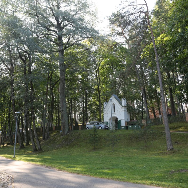 A white chapel surrounded by tall trees at the Roman Catholic cemetery in Druskininkai. A car is parked nearby on a grassy hill, with a paved path leading through the area.