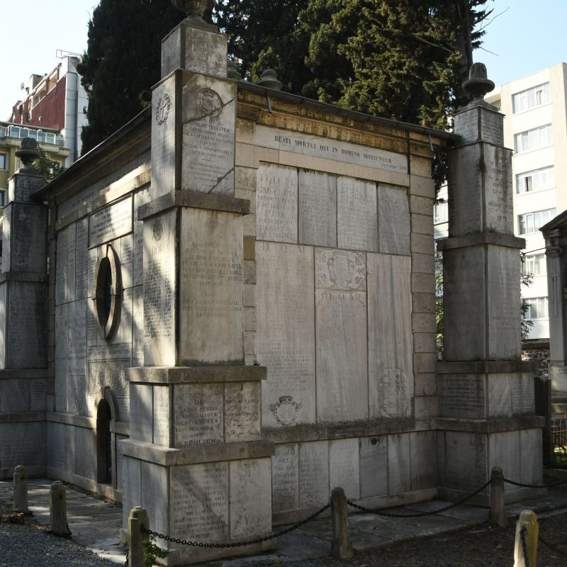 Rectangular tombstone with engraved uppercase inscription. The first two lines have slightly larger letters. Located in a cemetery with trees and buildings in the background.