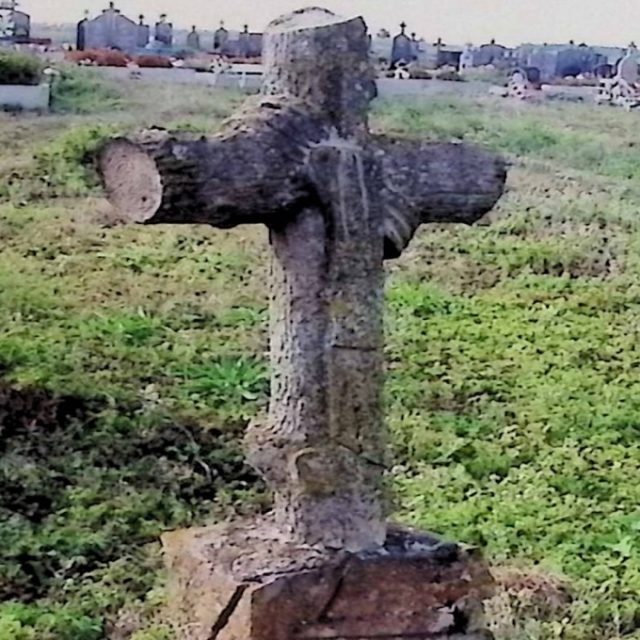 Pierre tombale d'Anastazja et Jan Rudnicki avec une croix stylisée comme un tronc d'arbre sur une base en forme de rocher. Un ruban sculpté est drapé sur les bras de la croix. Une plaque ressemblant à un parchemin avec une inscription est à l'avant.