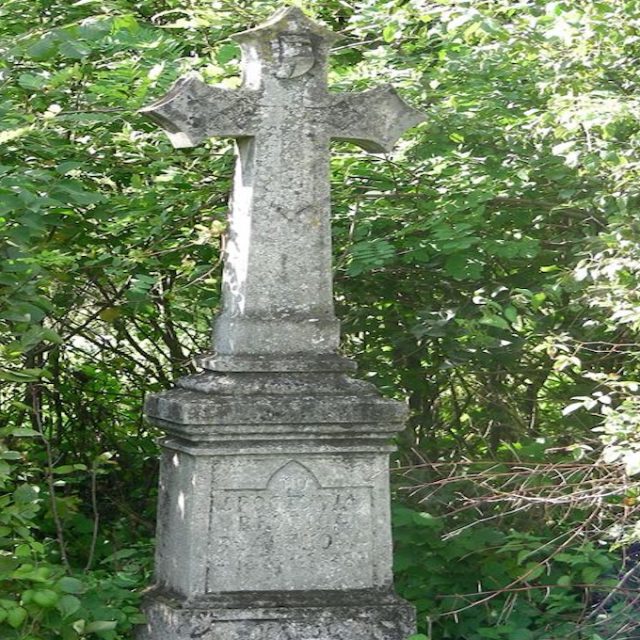 Tombe de Brygida et Karol Błaszkow avec une croix sur un piédestal à deux niveaux, entourée de verdure. La croix a une plus petite croix à l'intersection de ses bras et une inscription à la base.