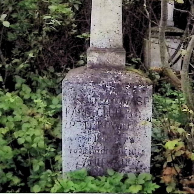 Tombe marquée par une croix sur un piédestal avec une inscription à l'avant. La croix est plus large en bas et repose sur un socle aux bords biseautés. Entourée de verdure.