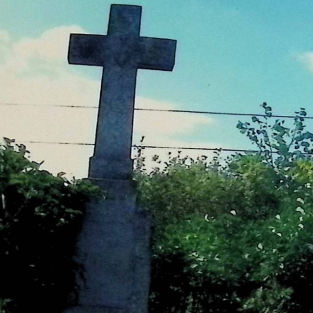 Надгробок з хрестом на трирівневому постаменті, оточений зеленню. Хрест прикрашений рельєфом палаючого серця з хрестом на перетині його рук.