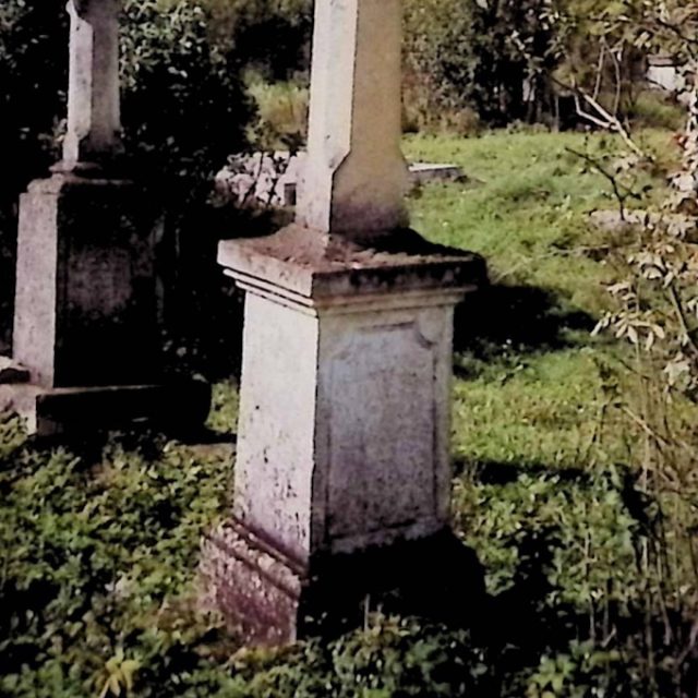 Pierre tombale avec une croix sur un piédestal à deux niveaux. La croix a des bras octogonaux avec des saillies. Le premier niveau a des bords profilés, le second est couvert d'une plaque plus large. Une inscription est gravée sur le panneau avant.