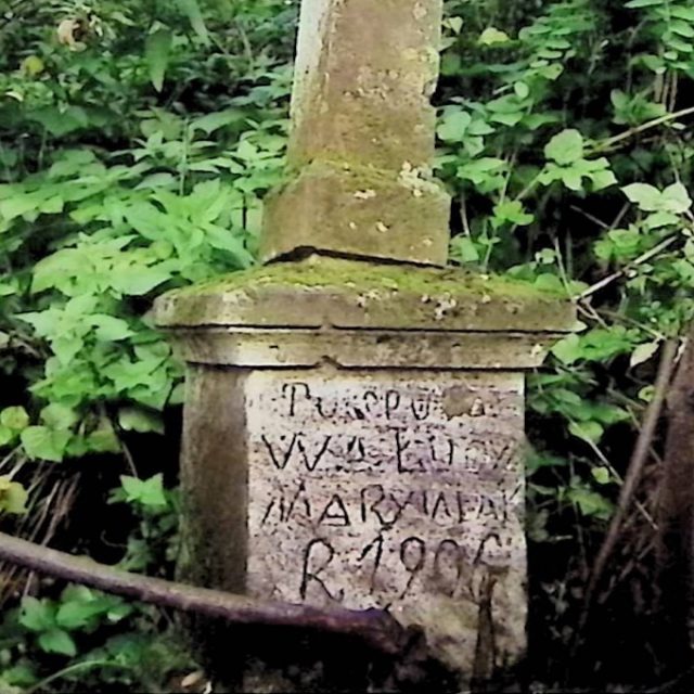 Tombe marquée par une croix simple sur un socle avec une inscription à l'avant. Le socle se transforme en toit à quatre pans. Entouré de verdure.