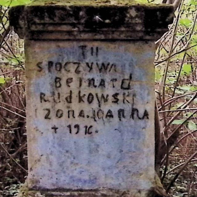 Gravestone of Bernard and Joanna Rudkowski with an inscription on a two-tiered pedestal, surrounded by vegetation. The cross is missing. The inscription reads: 'Here rests Bernard Rudkowski and wife Joanna +1916.'