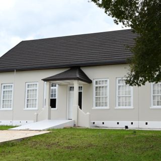 A white, single-storey building with a dark roof and lots of windows, surrounded by grass. The entrance has a small porch and steps leading up to it.