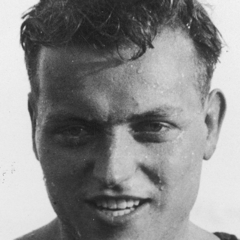 Close-up of a man with short hair, wearing a sleeveless top, smiling slightly. The image is in black and white.
