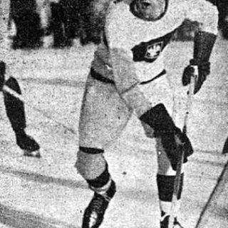 Un joueur de hockey en uniforme blanc avec un emblème polonais patine sur la glace, tenant un bâton de hockey. Des spectateurs sont visibles en arrière-plan.