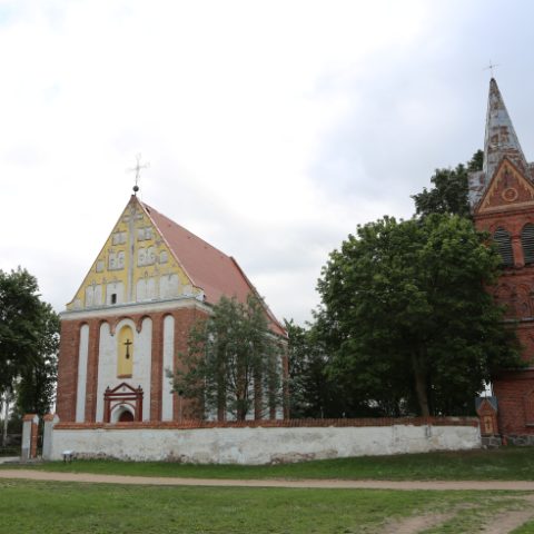 Gotycki kościół w Skorulach na Litwie z czerwoną ceglaną fasadą, trójkątnym frontonem i krzyżem na szczycie. Obok wysoka, czerwona ceglana dzwonnica. Kościół otaczają drzewa i biały tynkowany mur.