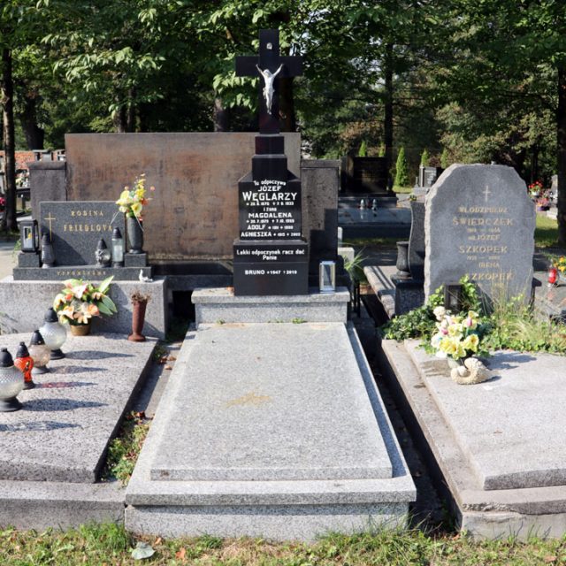 Tombe de la famille Węglarzy avec une pierre tombale ornée d'une croix sur un piédestal à deux niveaux. La croix porte une figure du Christ et un parchemin au-dessus. La tombe est entourée d'autres pierres tombales et de verdure.