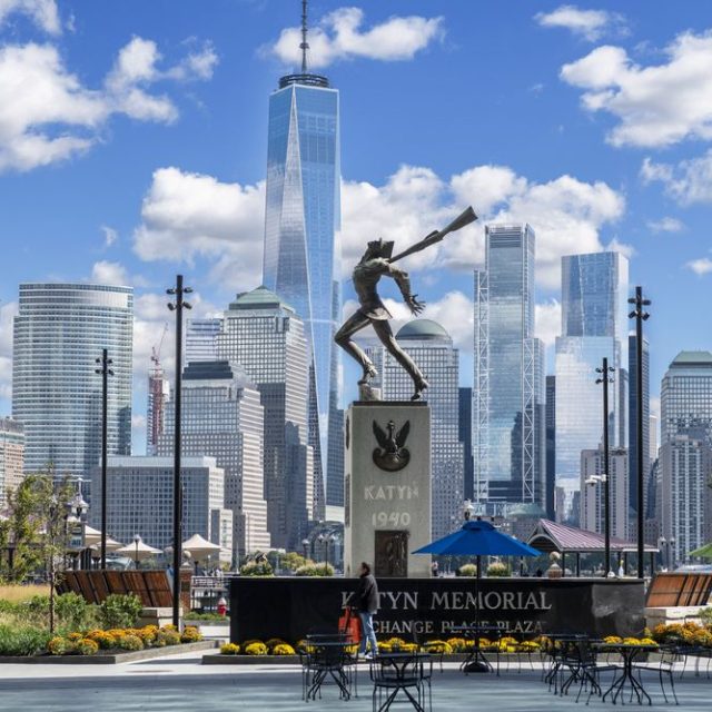 Pomnik Katyński w Jersey City z brązową statuą polskiego oficera na cokole, na tle panoramy Nowego Jorku. Na cokole widnieje napis 'Katyń 1940'.