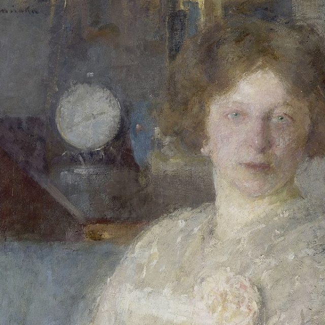 Portret kobiety o jasnych włosach i neutralnym wyrazie twarzy, w jasnej bluzce z kwiatowym detalem. Tło jest rozmyte z widocznym okrągłym obiektem.