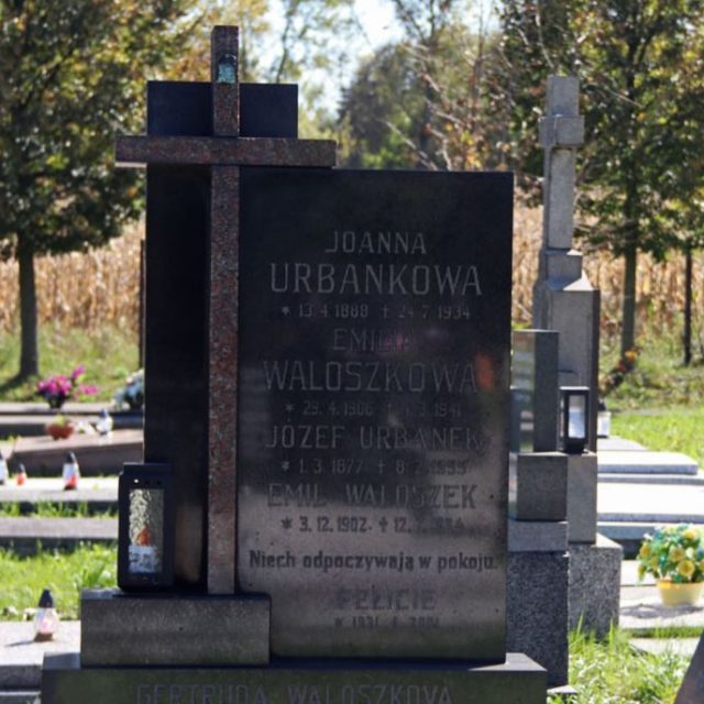 Pierre tombale des familles Waloszek et Urbanek avec inscriptions et une croix. Une petite lanterne est placée à la base. En arrière-plan, d'autres tombes et des arbres.