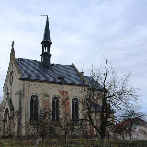 La chapelle publique de Młyniska, en Ukraine, construite en 1912, présente une structure en brique et pierre avec une haute flèche, des fenêtres cintrées et une façade vieillie. Des arbres sans feuilles entourent le bâtiment.