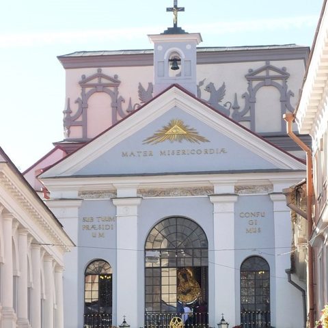 Fasada Kaplicy Ostrobramskiej w Wilnie z trójkątnym tympanonem z napisem 'Mater Misericordiae' i łukowatymi oknami. Kaplica otoczona klasycznymi kolumnami.