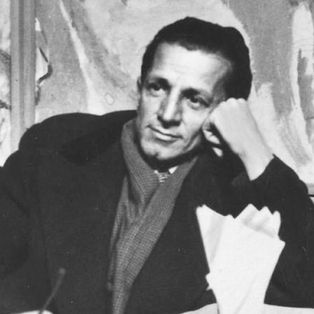 Photographie en noir et blanc d'un homme assis, la tête appuyée sur sa main, portant un manteau et une écharpe. Une œuvre d'art abstraite est visible en arrière-plan.