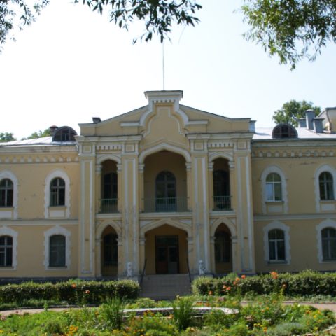 The neogothic palace in Priluki, Belarus, features a two-story structure with a central risalit, pointed arch windows, and corner turrets with crenellation. The garden in front is landscaped with flowers.