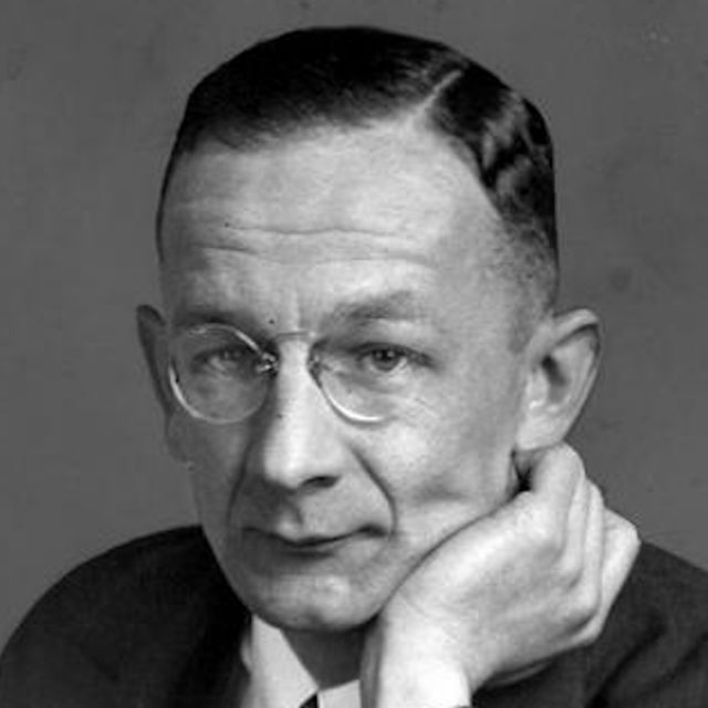 Portrait d'un homme avec des lunettes, appuyant son menton sur sa main, portant un costume et une cravate.