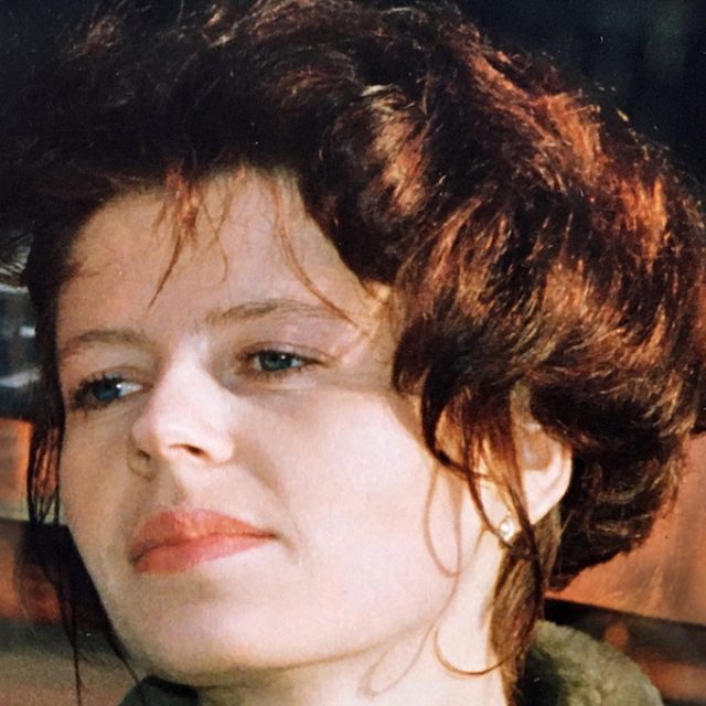 Portrait d'une femme aux cheveux bruns courts et ondulés, regardant sur le côté. Elle a une expression neutre et porte un haut foncé. L'arrière-plan est flou.