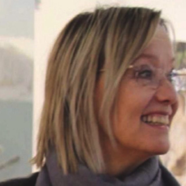 Une femme aux cheveux blonds mi-longs et lunettes sourit en regardant sur le côté.