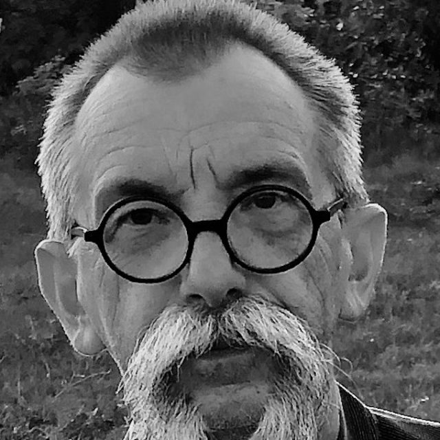 Czarno-biały portret mężczyzny w okularach i z wąsami, stojącego na zewnątrz z rozmytym tłem roślinnym.