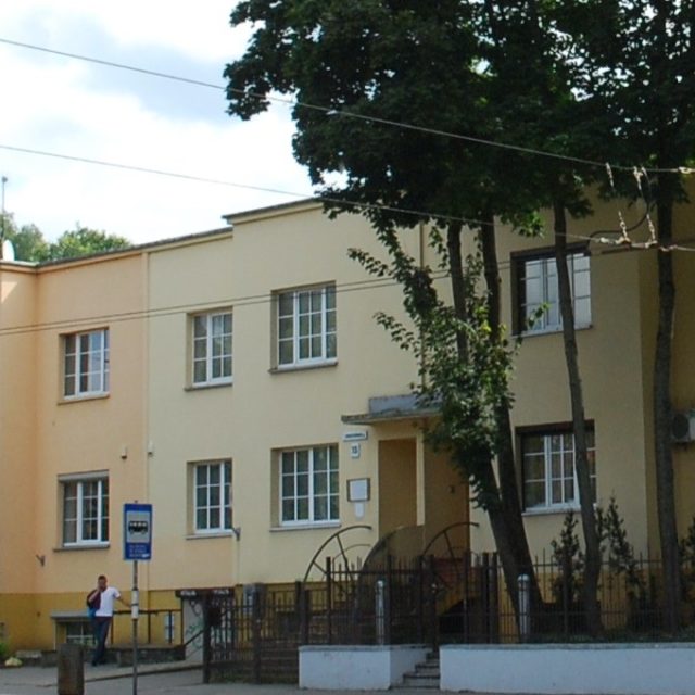 Dwupiętrowy budynek mieszkalny w Wilnie, część osiedla Spółdzielni Mieszkaniowej Ministerstwa Robót Publicznych. Fasada z dużymi oknami i płaskim dachem, z drzewami i ogrodzeniem z przodu.