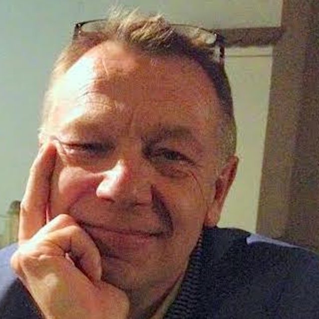 Osoba z krótkimi włosami i okularami na głowie, uśmiechająca się z ręką na policzku.