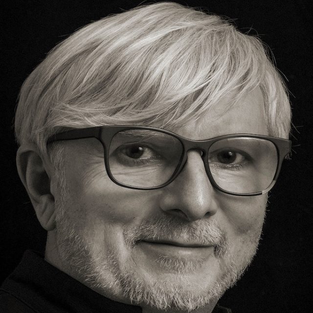 Portret mężczyzny w okularach i jasnych włosach, lekko uśmiechniętego, na ciemnym tle.
