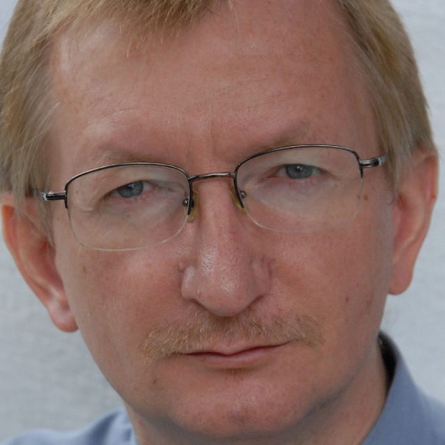 Portret mężczyzny w okularach i z wąsami, w niebieskiej koszuli, na jasnym tle.