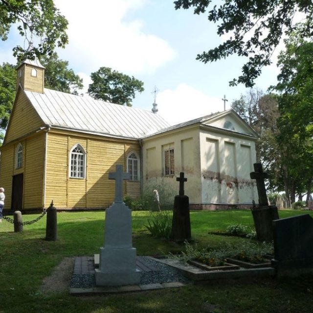 L'église en bois de la Transfiguration du Seigneur à Świętobrość, avec son toit métallique et son cimetière attenant. L'église possède des fenêtres en arc et un clocher. On aperçoit des pierres tombales au premier plan.
