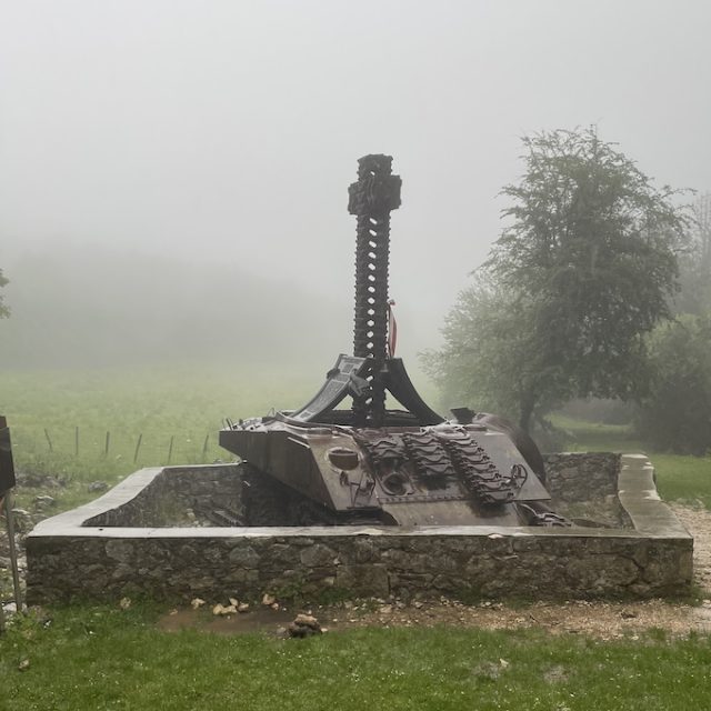 Фотографія пам'ятника танку «Султан» біля Монте-Кассіно. Пошкоджений корпус танка стоїть у невисокій кам'яній огорожі з високим хрестом із гусениць. Пам'ятник оточують трава, дерева та туман.