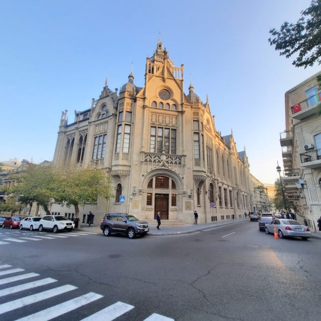 Fotografia pałacu Ismailija w Baku na rogu ulicy, z wysoką kamienną fasadą, smukłymi wieżami, arkadowym wejściem, zaparkowanymi samochodami i przejściem dla pieszych.