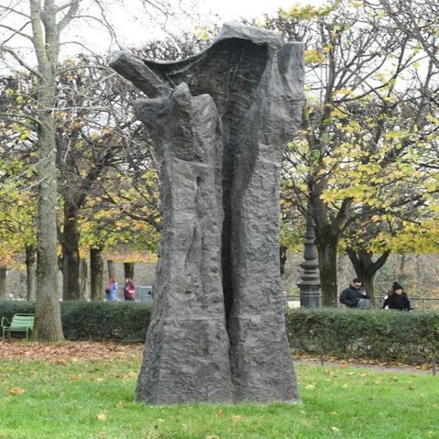 Bronze sculpture in the Tuileries Garden in Paris with a tall split form resembling a tree trunk or raised hand among grass and autumn trees.