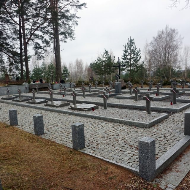 Military cemetery in Ławkiesy with rows of graves marked by metal crosses and stone slabs. Trees and a forested background are visible.