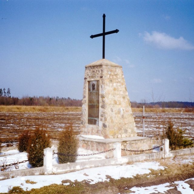 Кам'яний пам'ятник із хрестом на вершині, оточений невисокою огорожею та невеликими кущами, розташований на засніженому полі. Небо чисте, з кількома хмарами.