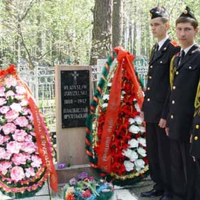 The grave of Wladyslaw Jaruzelski with a red sandstone plaque with his name and dates. Next to it are two floral wreaths with red ribbons. Nearby stand four young men in uniform.