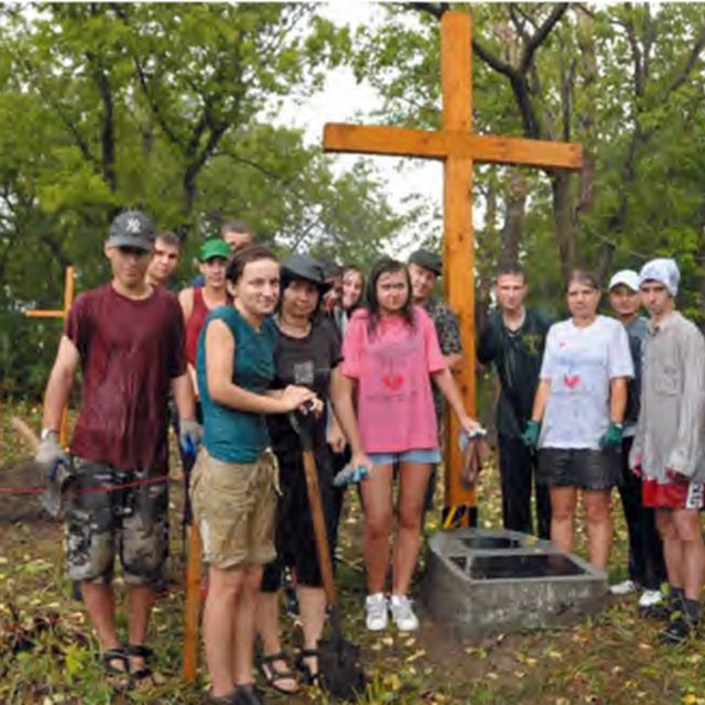 Grupa osób stoi wokół drewnianego krzyża na cmentarzu. Krzyż otaczają drzewa i kilka drewnianych znaczników. Ziemia pokryta jest liśćmi i trawą.