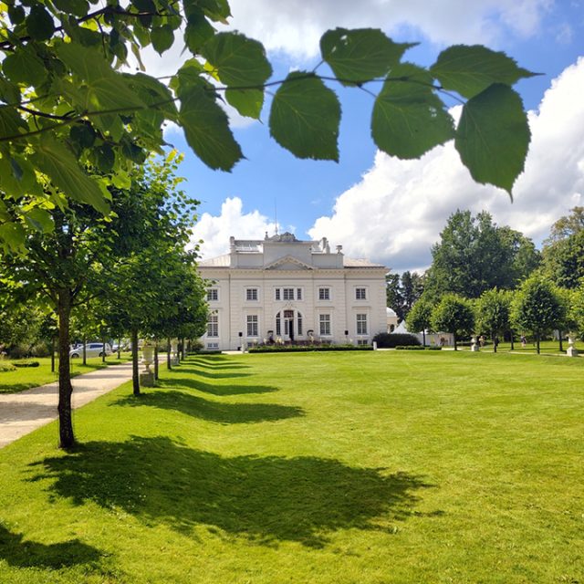 The Neoclassical Tyszkiewicz Palace in Zatrocz, Lithuania, with its white façade and two-storey structure, is surrounded by a landscaped park with trees and a lawn beneath a blue sky.