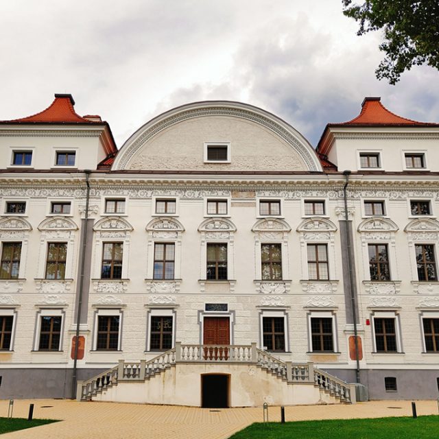 Fasada Pałacu Sapiehów na Antokolu w Wilnie, z symetrycznym układem, dekoracyjnymi elementami i centralnym wejściem ze schodami. Budynek ma białą elewację z czerwonymi akcentami dachowymi.