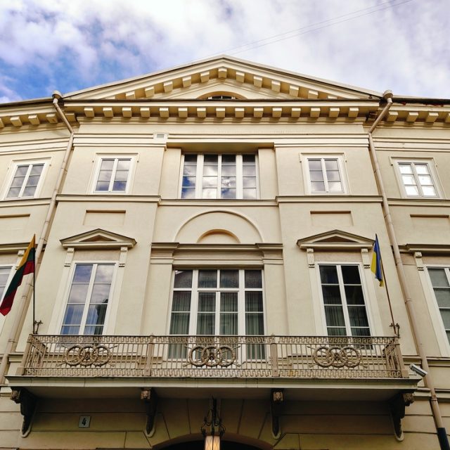 Fasada Pałacu Chodkiewiczów w Wilnie z dekoracyjnym balkonem i flagami. Budynek ma klasyczne elementy architektoniczne i symetryczny układ okien,  niebo pochmurne.