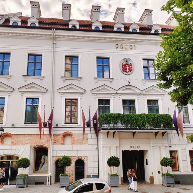 Façade du palais Pac à Vilnius sur la rue Wielka, avec une structure à trois étages, une entrée centrale, des colonnes toscanes et un balcon. Le bâtiment est décoré de drapeaux et de verdure.