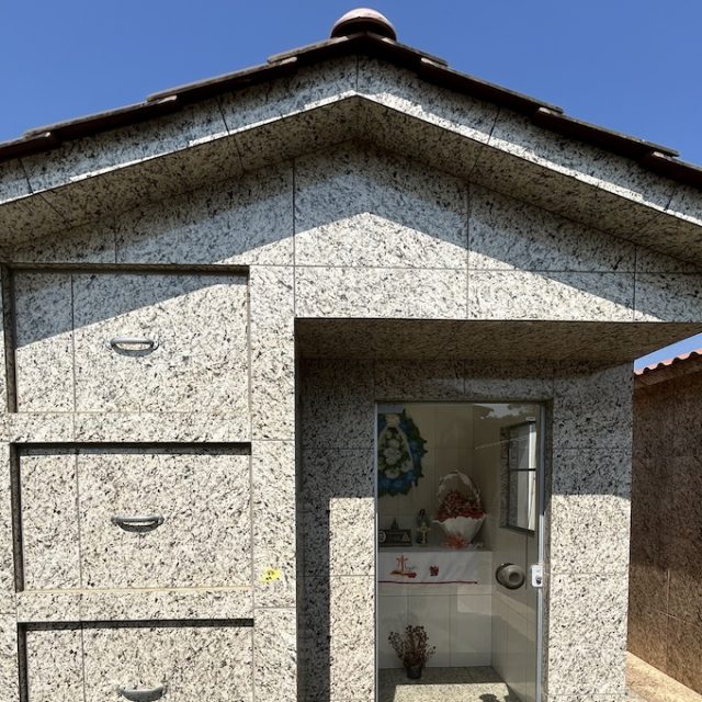 Petite chapelle en pierre avec un toit à pignon et une porte en verre. À l'intérieur, des objets religieux et une couronne de fleurs sont visibles.