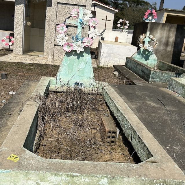Tombe d'Irena Michalska avec une pierre tombale turquoise ornée de fleurs artificielles roses et blanches. La tombe est entourée d'herbe sèche et située dans un cimetière avec d'autres tombes similaires.