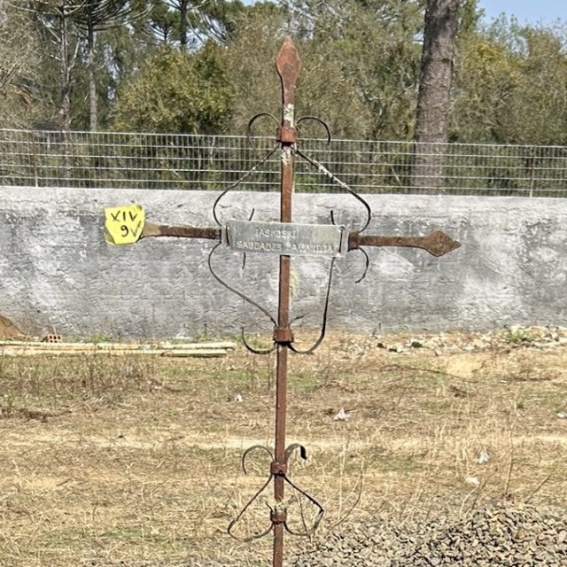 Rusted metal cross inscribed 'Jaskowski Saudades da Familia' in cemetery. Attached to one arm is a yellow marker with 'XIV 9V'. In the background, trees and a fence.