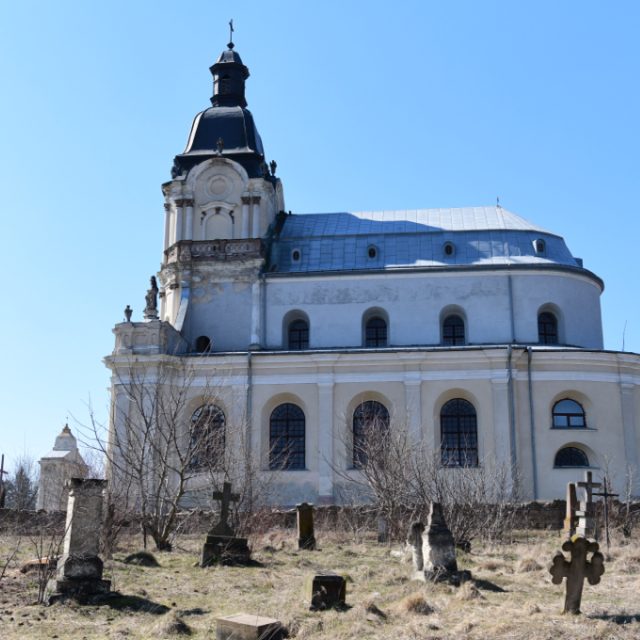 Side view of the Holy Trinity Church and Missionary Monastery in Mikulińce, showing baroque architecture with a dome and arched windows. In the foreground, old gravestones and crosses are visible.