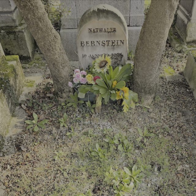 Nagrobek Natalii Bernstein z półokrągłym zakończeniem, z inskrypcją jej imienia i daty. Otoczony drzewami i kwiatami, kamień wkopany w ziemię.