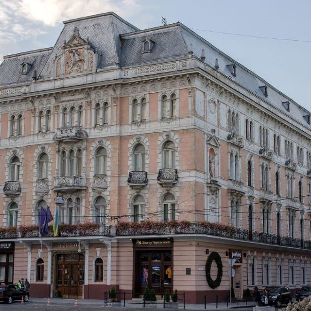 Hotel George we Lwowie, zabytkowy budynek z dachem mansardowym i ozdobną fasadą z balkonami i rzeźbami. Wejście flankują flagi, a ulica jest pełna zaparkowanych samochodów.